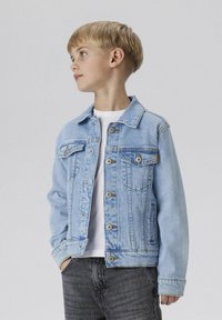Hellblaues Jeansjacke mit zwei Brusttaschen und Knopfleiste, kombiniert mit einem weißen Hemd und grauen Jeans. Glatte Textur und lockere Passform.
