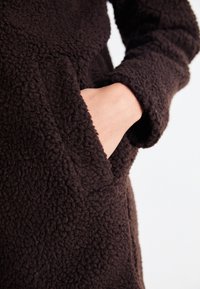 Manteau en polaire marron avec une surface texturée. Il dispose d'une poche avant et de longues manches, mettant en avant un matériau doux et confortable.