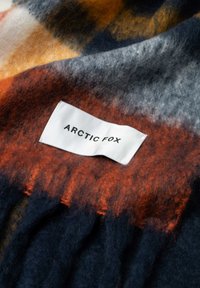 Couverture en laine avec un motif à carreaux orange, gris et bleu marine. Présente des bords à franges et une étiquette blanche avec "ARCTIC FOX" en texte noir.