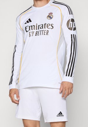 Man wearing white Real Madrid long-sleeve soccer jersey and shorts with black Adidas logos and sponsor branding on front and sleeves.