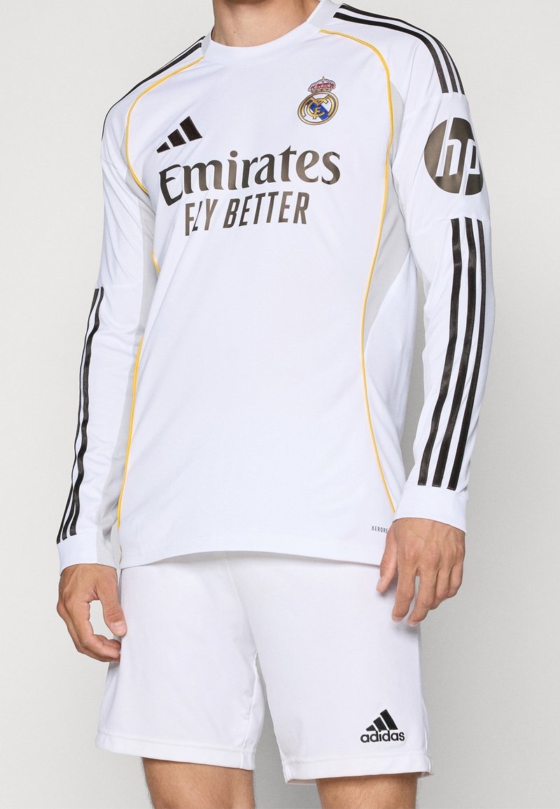 Homme portant un maillot de football Real Madrid à manches longues blanc et un short avec logos Adidas noirs et marquages des sponsors sur le devant et les manches.