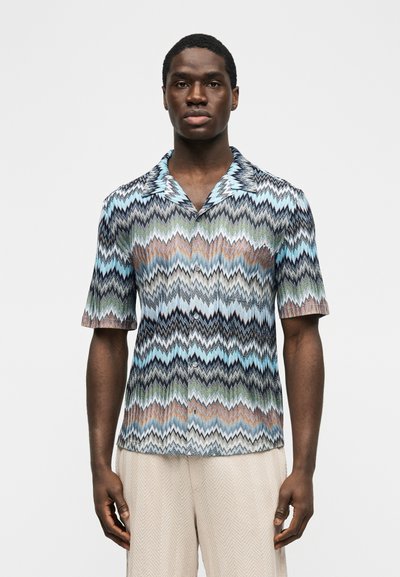 Άνδρας με πουκάμισο με κοντά μανίκια και πολύχρωμο ζιγκ-ζαγκ μοτίβο και ανοιχτό μπεζ υφασμάτινο παντελόνι, σε απλό φόντο.