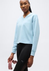 Γυναίκα που φοράει ανοιχτό μπλε φούτερ και μαύρο κολάν Puma, κρατώντας μια ροζ ελαστική ταινία με το δεξί της χέρι, στέκεται μπροστά σε ένα απλό λευκό φόντο.