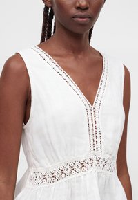 Gros plan d'une personne portant une robe blanche sans manches avec une finition en dentelle le long du décolleté en V et de la taille.