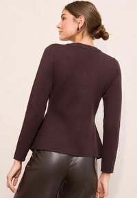 Tumši brūns garām piedurknēm tops ar pieguļošu virsdaļu un izplešamu apakšdaļu, izgatavots no teksturēta auduma. Nēsāts kopā ar spīdīgiem brūniem ādas biksēm.