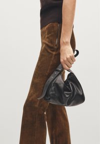 Black leather handbag with soft, crinkled texture and a textured strap held in one hand, paired with brown suede wide-leg trousers.