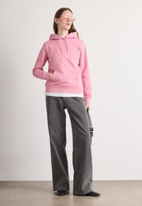 Sudadera rosa con un bolsillo frontal y capucha con cordón, vestida sobre una camisa blanca, combinada con pantalones de pierna ancha gris oscuro y zapatos negros.