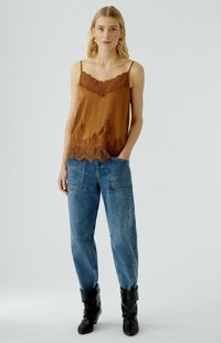 Blonde vrouw in een bruine cami met kant, losse blauwe jeans met grote zakken en zwarte slouchy laarzen, staand met één hand in haar zak.