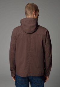 Chaqueta ligera marrón con capucha, con un diseño simple y puños elásticos, llevada sobre unos jeans azules, vista por la parte de atrás.