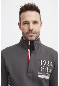 Felpa grigia con colletto zip, caratterizzata da accenti rossi e bianchi, testo ricamato e una texture liscia. Logo notevole sul petto.