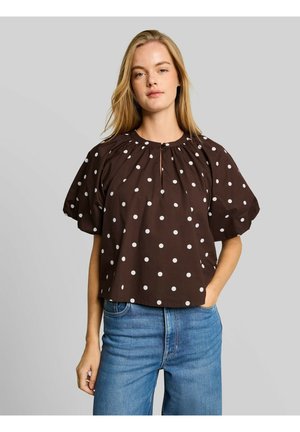 Giovane donna che indossa una camicetta marrone con pois bianchi e jeans blu, in piedi con una mano in tasca davanti a uno sfondo grigio.