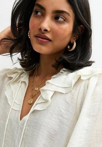 Witte, gerimpelde blouse van lichtgewicht stof. Gelaagde gouden kettingen en gedraaide ringoorbellen voegen accenten toe aan de uitstraling. Gladde, zachte textuur.