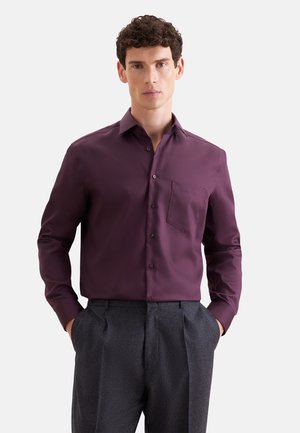 Homme portant une chemise boutonnée à manches longues violet foncé avec une poche poitrine et un pantalon plissé gris foncé, mains dans les poches.