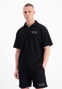 Zwart gestructureerd poloshirt met korte mouwen en een V-hals, voorzien van een borduring op de borst, in combinatie met bijpassende zwarte gestructureerde shorts.