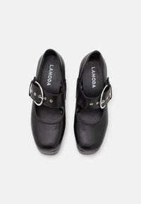 Svarta Mary Jane-skor med ett brett band och silverfärgad spänne, glänsande yta, rund tå och upphöjd platåsål. Varumärken "LAMODA" synligt inuti.