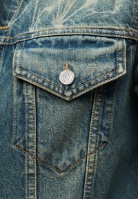 Κοντινή λήψη τσέπης μπλε τζιν μπουφάν με τριγωνικό καπάκι ασφαλισμένο με ασημένιο κουμπί με χαραγμένο προφίλ.