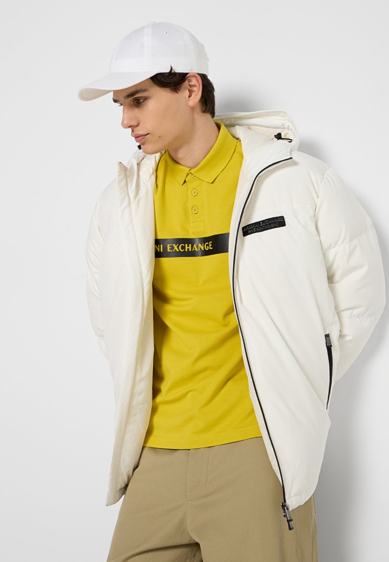Giacca a vento bianca con cappuccio e zip nera. Indossata sopra una polo gialla con un logo nero. Pantaloni grigi chiari. Cappellino bianco.