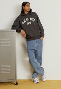 Sudadera gris con capucha que lleva el texto "NEW BALANCE 1906", combinada con jeans sueltos de color azul claro y zapatillas blancas. Taquilla metálica de fondo.