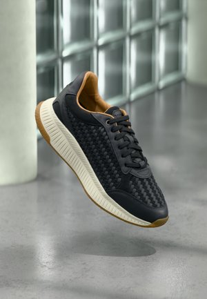 Scarpa da ginnastica nera con motivo intrecciato, suola bianca testurizzata e interno marrone, sospesa sopra un pavimento grigio vicino a una parete in vetrocemento.