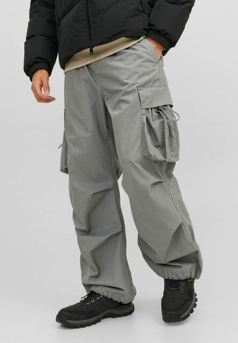 Pantalon cargo gris à coupe ample, avec plusieurs poches latérales, des poignets élastiques et un tissu léger. Porté avec des chaussures noires.