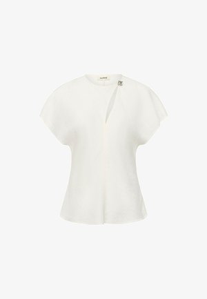 Blusa bianca a maniche corte con una texture sottile, caratterizzata da una piccola fibbia decorativa argentata vicino allo scollo rotondo e una fessura frontale.