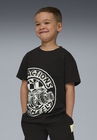 Lächelnder Junge mit kurzen Haaren, der ein schwarzes T-Shirt mit einem großen Monstertruck-Motiv und Text trägt, die Hände in den Taschen, vor einem grauen Hintergrund.