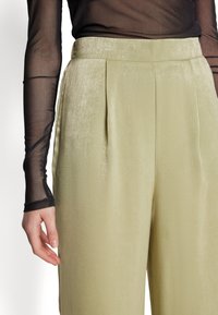 Pleated trousers in light green fabric with a smooth texture. A sheer black long-sleeve top partially visible on the left.
