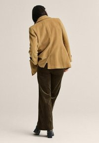 Ruskea villasekoitebleiseri, jossa on jämäkät olkapäät, takakslit ja joka on yhdistetty vihreisiin leveälahkeisiin puuvillafarkkuihin sekä mustiin nilkkureihin.