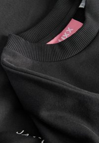 Černá mikina se žebrovaným límcem a manžetami. Vnitřní část límce obsahuje růžovou etiketu. Tkanina je hladká s mírným leskem a má minimalistický design.