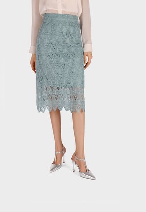 Femme portant une jupe en dentelle bleue jusqu'aux genoux, un chemisier blanc transparent et des chaussures à talons hauts argentées avec bride en T, debout sur un fond uni.