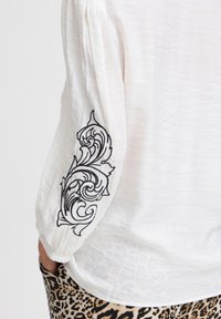 Hvid langærmet bluse med sort broderet blomsterdesign på ærmet, med tekstureret stof og bløde samlede manchetter.