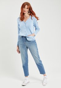 Lichtblauwe cardigan met een V-hals, knoopsluiting en lange mouwen, gecombineerd met high-waisted lichtblauwe spijkerbroeken en witte sneakers.