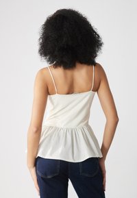 Bílý peplum top s tenkými ramínky, hladkým materiálem a nabraným pasem; nošený s tmavě modrými džíny. Pohled zezadu.