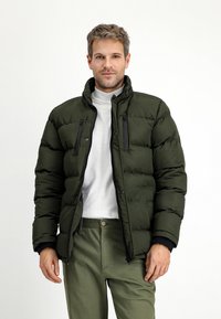 Donkergroene pufferjack met een hoge kraag, ritszakken op de borst en een gequilte textuur. Gecombineerd met een lichtgrijze coltrui.