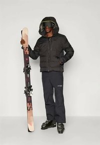 Síruha, amely fekete puffos dzsekiből kapucnival, sötét nadrágból és sícipőből áll. Egy sílécet tart kötésekkel; a síszemüveg az arcon van.