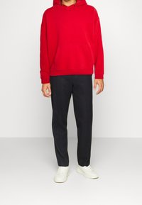 Czerwona bluza z kapturem oraz kieszenią z przodu, w zestawieniu z ciemnymi, zwężanymi spodniami i białymi sneakerami. Prosty design i gładka tekstura.