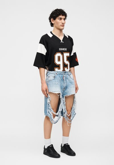 Νεαρό άτομο που φοράει μαύρη αθλητική φανέλα με το "95" και πολύ σκισμένα, φθαρμένα τζιν σορτς στέκεται μπροστά σε ένα απλό φόντο.