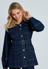 Chaqueta de mezclilla en azul oscuro, con cuello tipo solapa, bolsillos delanteros, botones dorados y una cintura ceñida con un cinturón que tiene una hebilla rectangular.