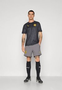 Puma-Fußballtrikot mit schwarzem, gemustertem Kurzarmshirt und grauen Shorts. Enthält gelbes Logo und Akzente, kombiniert mit schwarzen Socken und Schuhen.