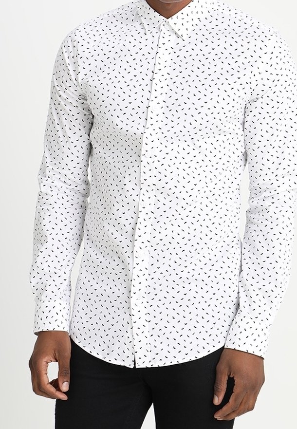 Hombre vestido con una camisa blanca ajustada de botones con pequeños patrones de hojas negras y pantalones negros, de pie contra un fondo blanco.