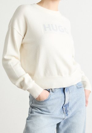 Henkilö, joka pukeutunut kermaanväriseen neulepaitaan, jossa lukee "HUGO", ja vaaleansinisiin farkkuihin, kädet taskuissa, yksinkertaista vaaleaa taustaa vasten.