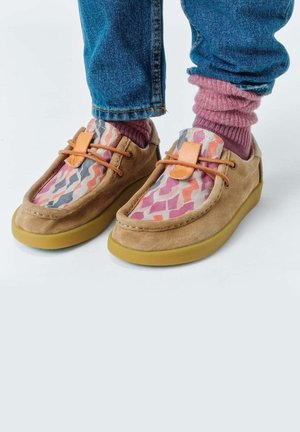 Pies con zapatos casuales de ante color canela con partes superiores con patrones multicolor y calcetines rosas bajo vaqueros azules sobre un fondo claro.