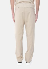 Pantaloni beige su misura con una vestibilità rilassata, texture liscia e una piega sottile sul retro, abbinati a sneakers bianche.