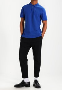 Man i blå polo skjorta, svarta byxor, vita strumpor och svarta dress skor, står med händerna i fickorna mot en vit bakgrund.