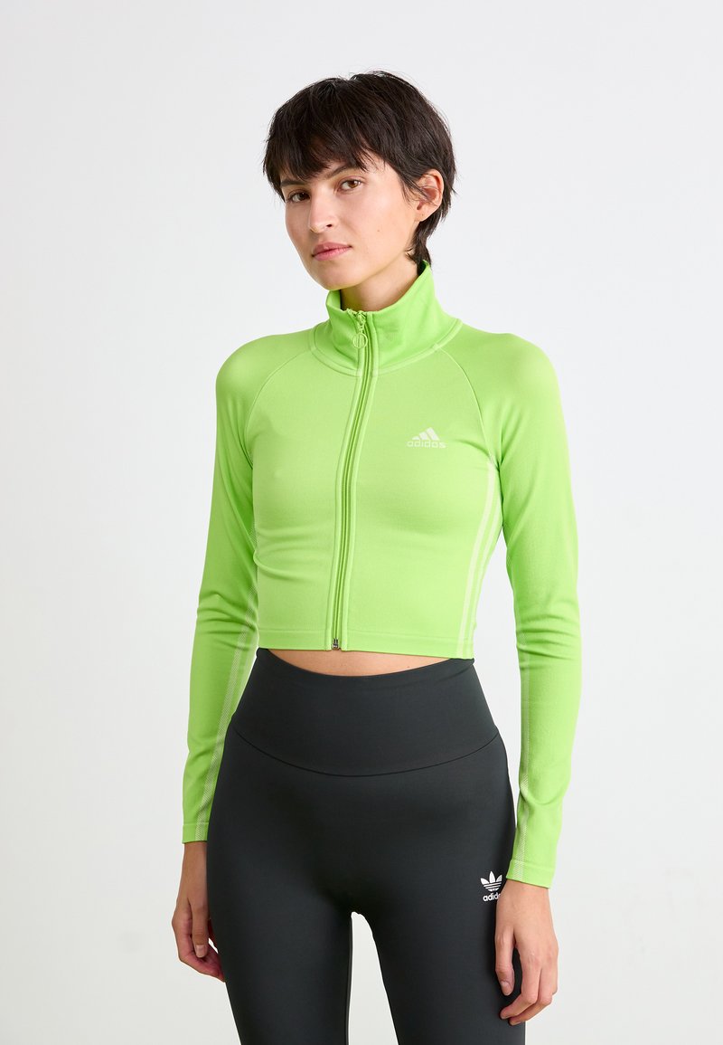 Kvinne i neon grønn cropped zip-up jakke og høy midje svart leggings med Adidas logoer på brystet og benet.