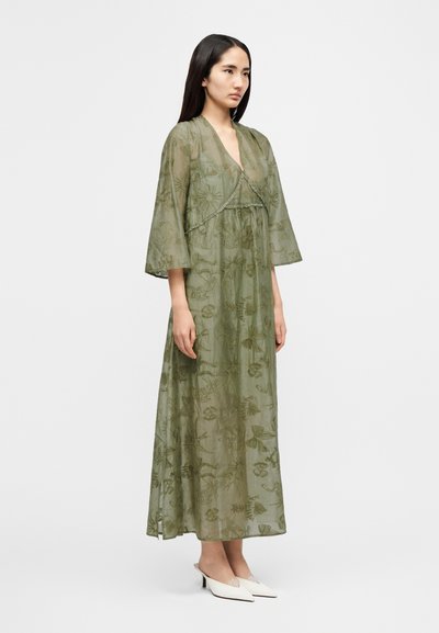 Μακρύ, ρέον πράσινο φόρεμα από διαφανές ύφασμα με κεντήματα λουλουδιών. Διαθέτει φαρδιές μανίκες και ξεχωριστό υποστρώμα. Συνδυάζεται με λευκά mule παπούτσια.