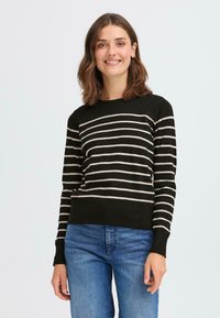 Maglione a righe nero e beige con collo rotondo, maniche lunghe e orlo a coste. Indossato sopra jeans blu. Tessuto morbido e design aderente.
