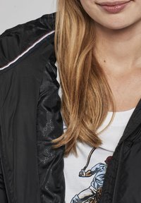 Czarna, lekka kurtka o błyszczącej fakturze, z suwakiem i kontrastowymi paskami na ramionach. Pod spodem biała koszula z graficznym wzorem.