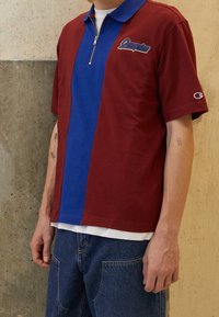 Personne portant un polo à manches courtes rouge et bleu avec une fermeture éclair, debout devant un mur en béton beige.