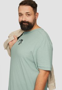 Lichtgroene katoenen t-shirt met korte mouwen, met een grafisch ontwerp op de borst. Een beige jas die over de schouder is gedrapeerd voegt detail toe.